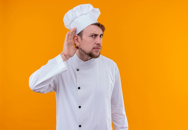 Um jovem chef barbudo em uniforme branco tentando ouvir algo em uma parede laranja