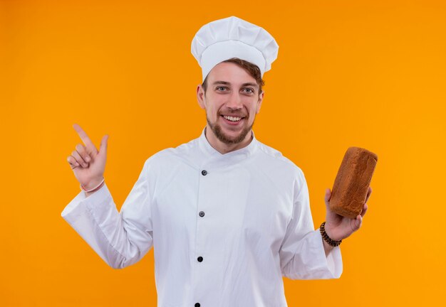 Foto grátis um jovem chef barbudo feliz com uniforme branco apontando para cima com o dedo indicador enquanto segura um pedaço de pão em uma parede laranja