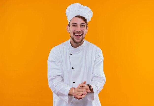 Um jovem chef barbudo feliz em uniforme branco, sorrindo e segurando as mãos juntas em uma parede laranja