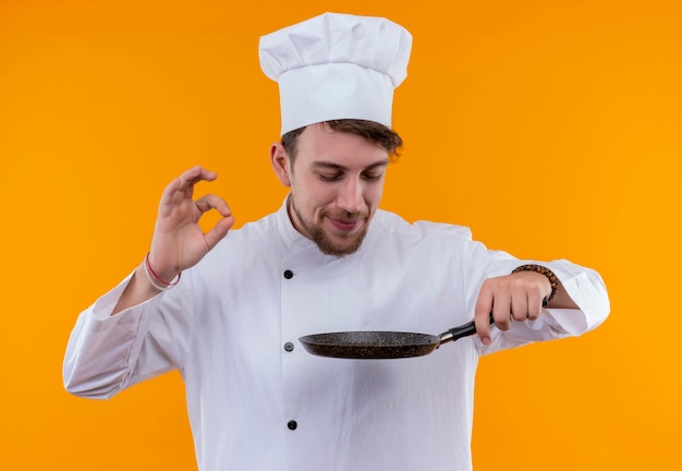 Foto grátis um jovem chef barbudo satisfeito, de uniforme branco, cheirando a frigideira e mostrando um saboroso gesto de ok em uma parede laranja