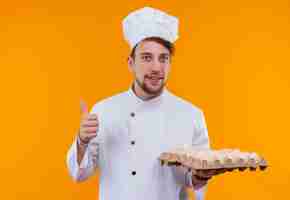 Foto grátis um jovem chef barbudo satisfeito vestindo uniforme branco de cozinheiro e chapéu segurando uma caixa de ovos enquanto olha para uma parede laranja