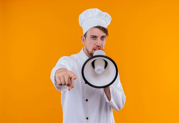 Um jovem chef barbudo sério, de uniforme branco, falando através do megafone enquanto olha para uma parede laranja