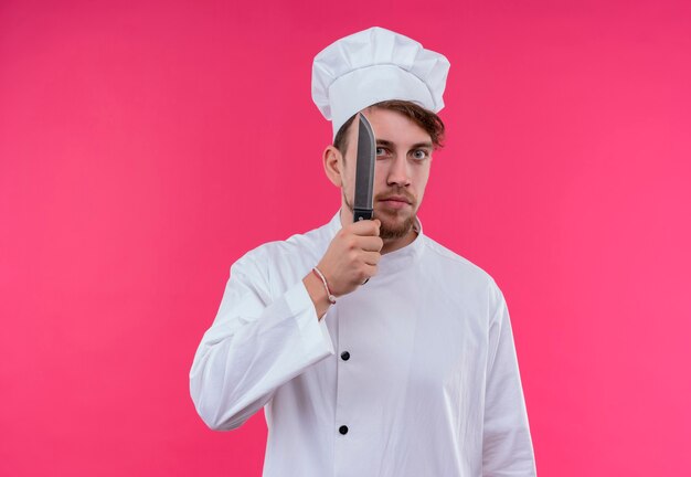 Um jovem chef barbudo sério, de uniforme branco, segurando uma faca afiada enquanto olha para uma parede rosa