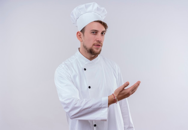 Foto grátis um jovem chef barbudo sério vestindo uniforme branco de fogão e chapéu convidativo a vir com a mão enquanto olha para uma parede branca