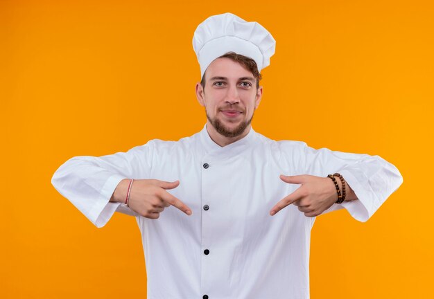 Foto grátis um jovem chef barbudo sorridente com uniforme branco apontando para baixo com o dedo indicador enquanto olha para uma parede laranja