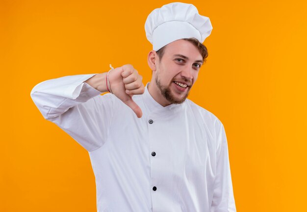 Um jovem chef barbudo sorridente de uniforme branco mostrando os polegares para baixo enquanto olha para uma parede laranja