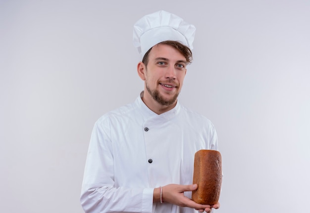 Foto grátis um jovem chef barbudo sorridente, vestindo uniforme branco de fogão e chapéu, segurando um pedaço de pão enquanto olha para uma parede branca