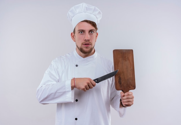 Um jovem chef barbudo vestindo uniforme branco de fogão e chapéu apontando para a placa de madeira da cozinha com uma faca enquanto olha para uma parede branca