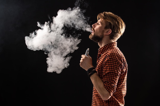 Um jovem com barba e um penteado elegante em uma camisa, fumando um cigarro, uma víbora, um quarto, um estúdio, fumaça, prazer. fundo preto