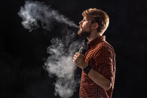 Um jovem com barba e um penteado elegante em uma camisa, fumando um cigarro, uma víbora, um quarto, um estúdio, fumaça, prazer. Fundo preto