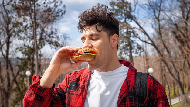 Foto grátis um jovem de camisa vermelha comendo um hambúrguer em um parque, árvores nuas