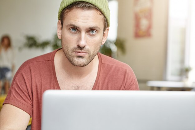Um jovem de olhos azuis e barba parece confiante enquanto se senta em frente a um laptop aberto, verifica e-mail ou navega em redes sociais online