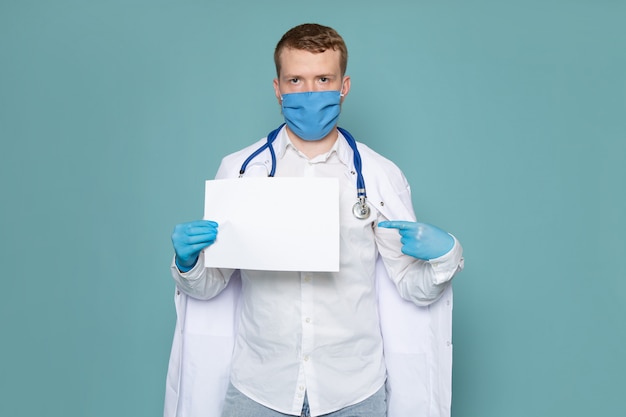 Um jovem de vista frontal na camisa branca e luvas azuis com máscara azul com pedaço de papel no espaço azul