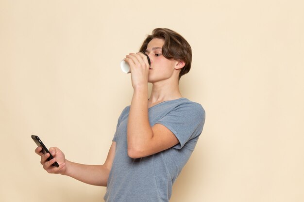 Um jovem do sexo masculino com camiseta cinza, bebendo café e usando um telefone