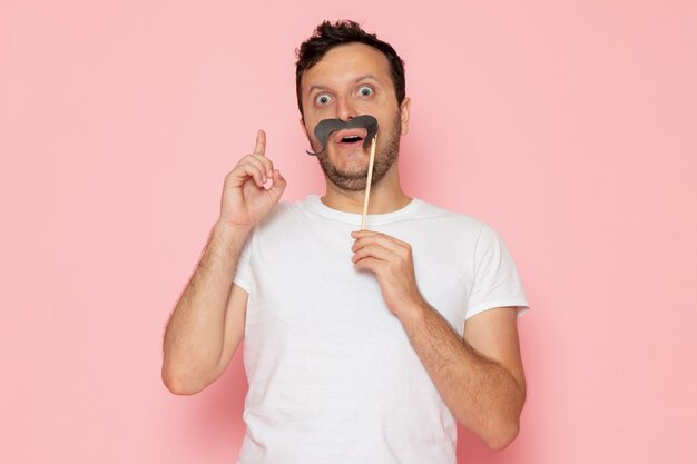 Um jovem do sexo masculino com uma camiseta branca segurando um bigode falso na mesa rosa homem cor de emoção pose