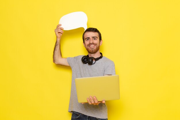 Um jovem do sexo masculino com uma camiseta cinza segurando uma placa branca com um sorriso na parede amarela homem expressão emoção cor modelo