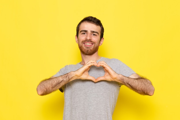 Um jovem do sexo masculino em uma camiseta cinza sorrindo e mostrando um sinal de amor na parede amarela.