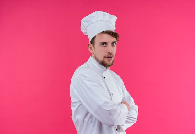 Um jovem e bonito chef barbudo em uniforme branco de mãos postas enquanto olha para uma parede rosa