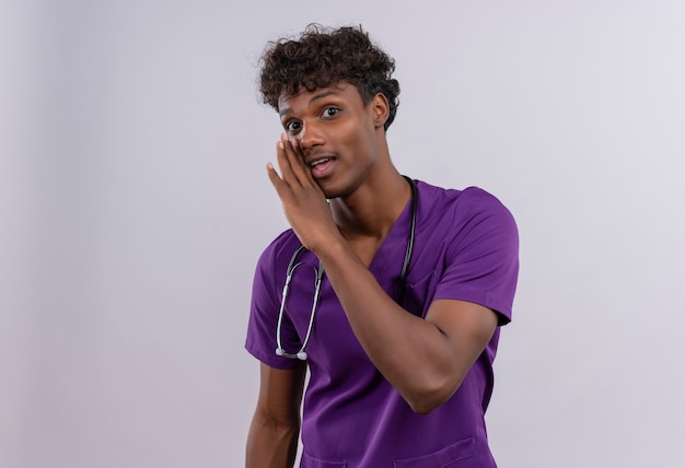 Um jovem e bonito médico de pele escura com cabelo encaracolado e uniforme violeta com estetoscópio tentando dizer algo