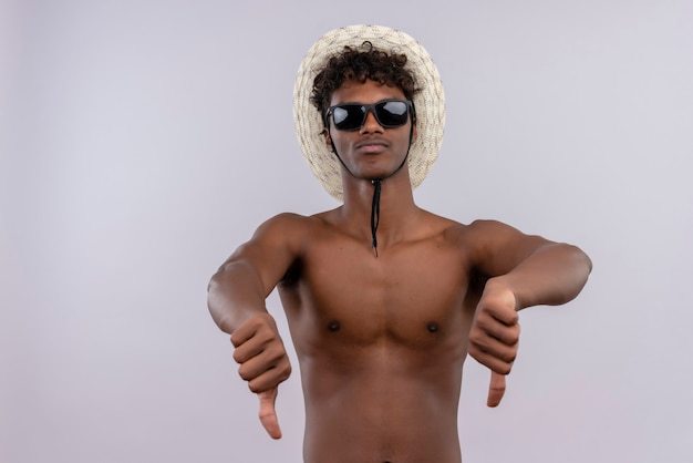 Um jovem e confiante homem bonito de pele escura com cabelo encaracolado usando chapéu de sol e óculos escuros mostrando os polegares para baixo