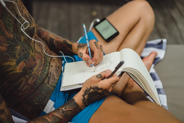 um jovem em tatuagens usando fones de ouvido ouve música e desenha em um notebook.