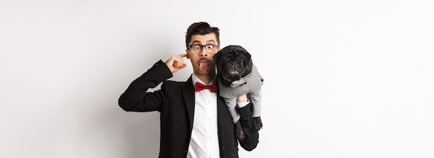 Foto grátis um jovem engraçado em traje de festa mostrando a língua e segurando um lindo pug preto no ombro comemorando com