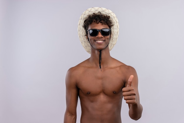 Um jovem feliz, bonito, de pele escura com cabelo encaracolado, usando chapéu de sol e óculos escuros mostrando os polegares para cima.