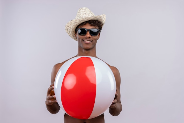 Um jovem feliz, bonito, de pele escura com cabelo encaracolado, usando chapéu de sol e óculos escuros, segurando uma bola inflável