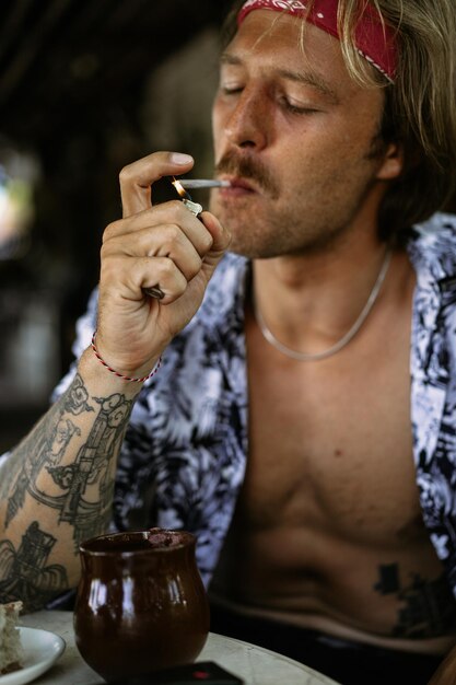 Um jovem hippie bonito em uma bandana vermelha com um torso nu rola um cigarro de tabaco em um café em Bali.