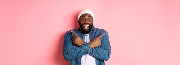 Foto grátis um jovem hipster africano-americano feliz apontando os dedos para o lado, sorrindo e mostrando duas opções