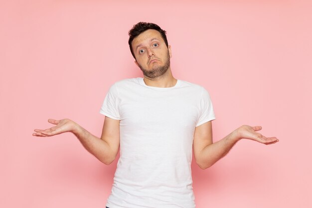 Um jovem homem de frente para uma camiseta branca e calça jeans posando com uma expressão confusa na mesa rosa.