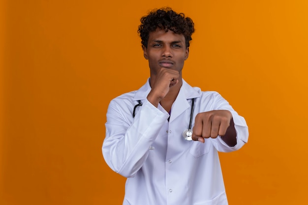 Foto grátis um jovem médico bonito, de pele escura, cabelo encaracolado, jaleco branco e estetoscópio, levantando as mãos com os punhos cerrados