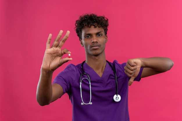 Um jovem médico bonito, de pele escura, com cabelo encaracolado, chateado, usando uniforme violeta com estetoscópio mostrando os polegares para baixo enquanto segura a pílula