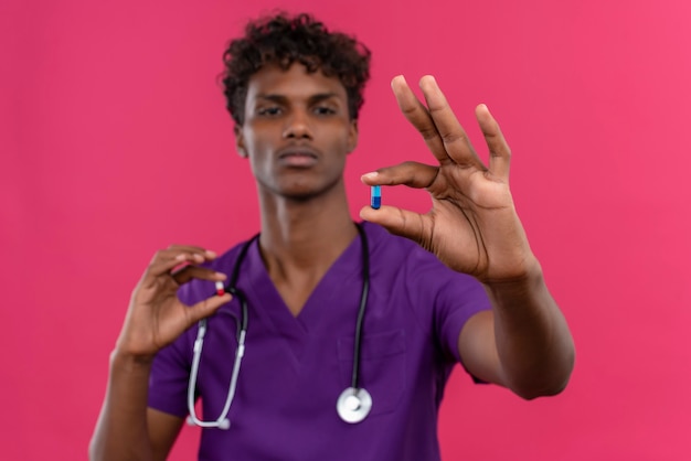 Foto grátis um jovem médico bonito, de pele escura com cabelo encaracolado, usando uniforme violeta com estetoscópio olhando para comprimidos