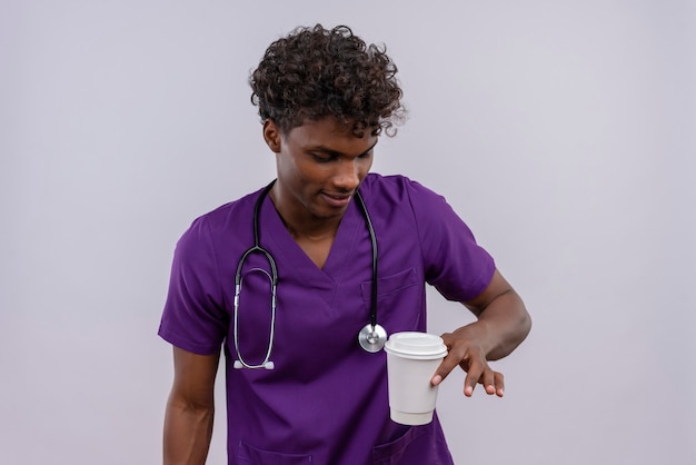 Um jovem médico bonito, de pele escura com cabelo encaracolado, usando uniforme violeta com estetoscópio olhando para um copo de papel de café