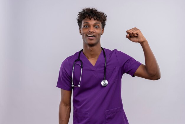 Um jovem médico de pele escura, animado e bonito, com cabelo encaracolado, usando uniforme violeta com estetoscópio olhando para seu smartphone com o punho cerrado