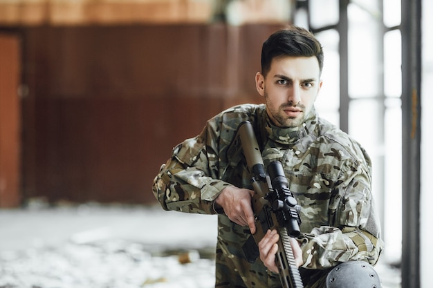 Um jovem soldado militar está sentado com um grande rifle nas mãos, perto da janela de um prédio desabado