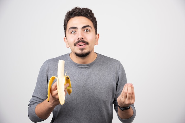 Um jovem sorridente comendo uma banana sobre uma parede branca.