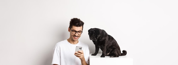 Um jovem sorridente usando smartphone e sentado perto do dono do cão pug verificando fotos no celular wh