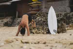 Foto grátis um jovem surfista bonito na costa do oceano está fazendo um aquecimento antes de surfar. exercícios antes do esporte, alongamento antes do surf.