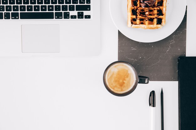 Um laptop aberto; waffle; xícara de café; lápis; caneta e laptop aberto na mesa branca