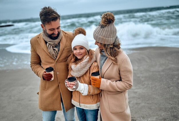 Um lindo casal e sua encantadora filha caminham à beira-mar e bebem café, vestidos com roupas quentes.