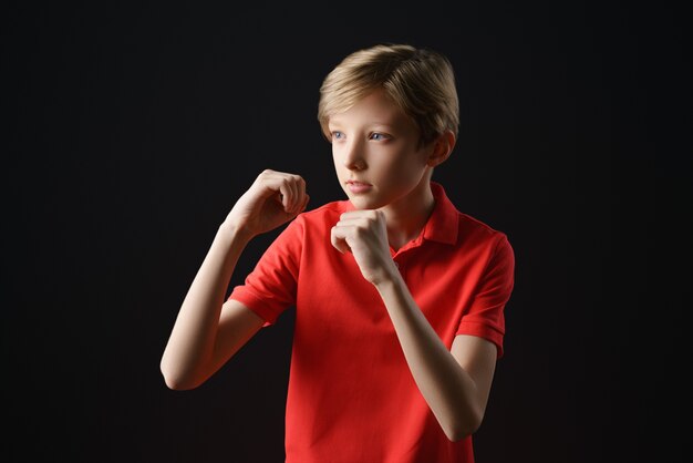 Um menino com uma camiseta vermelha com um corte de cabelo curto em um fundo preto segura as mãos como no boxe, uma pose de proteção