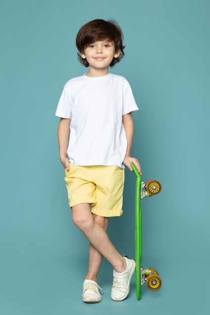 Um menino de criança bonito vista frontal em camiseta branca e calça jeans amarela, segurando o skate verde no chão azul