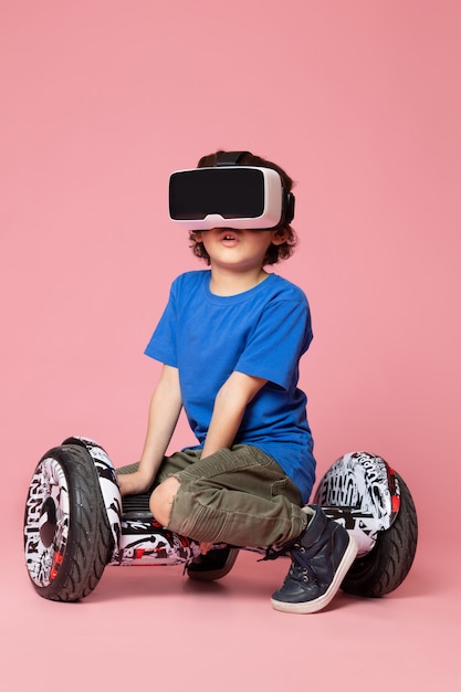 Um menino de criança bonito vista frontal em t-shirt azul jogando vr em segway no espaço rosa