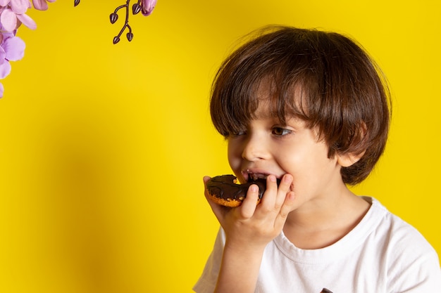 Foto grátis um menino de criança vista frontal comendo rosquinhas com chocolate no espaço amarelo