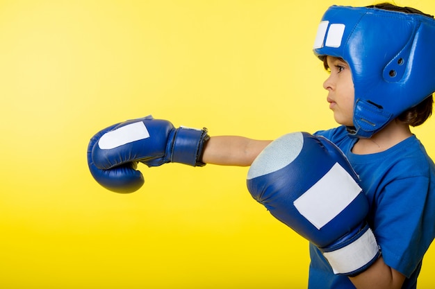 Um menino de criança vista frontal em luvas azuis e capacete azul boxe na parede amarela