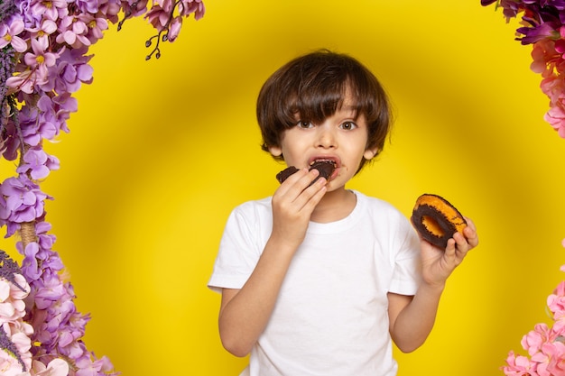 Foto grátis um menino de vista frontal comendo rosquinhas de chocolate em camiseta branca no espaço amarelo