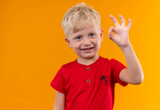 Um menino doce e fofo com cabelo loiro e olhos azuis, vestindo uma camiseta vermelha mostrando um gesto de ok com a mão