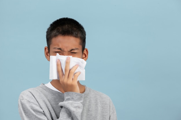 Um menino está espirrando nos tecidos e se sentindo doente na parede azul.